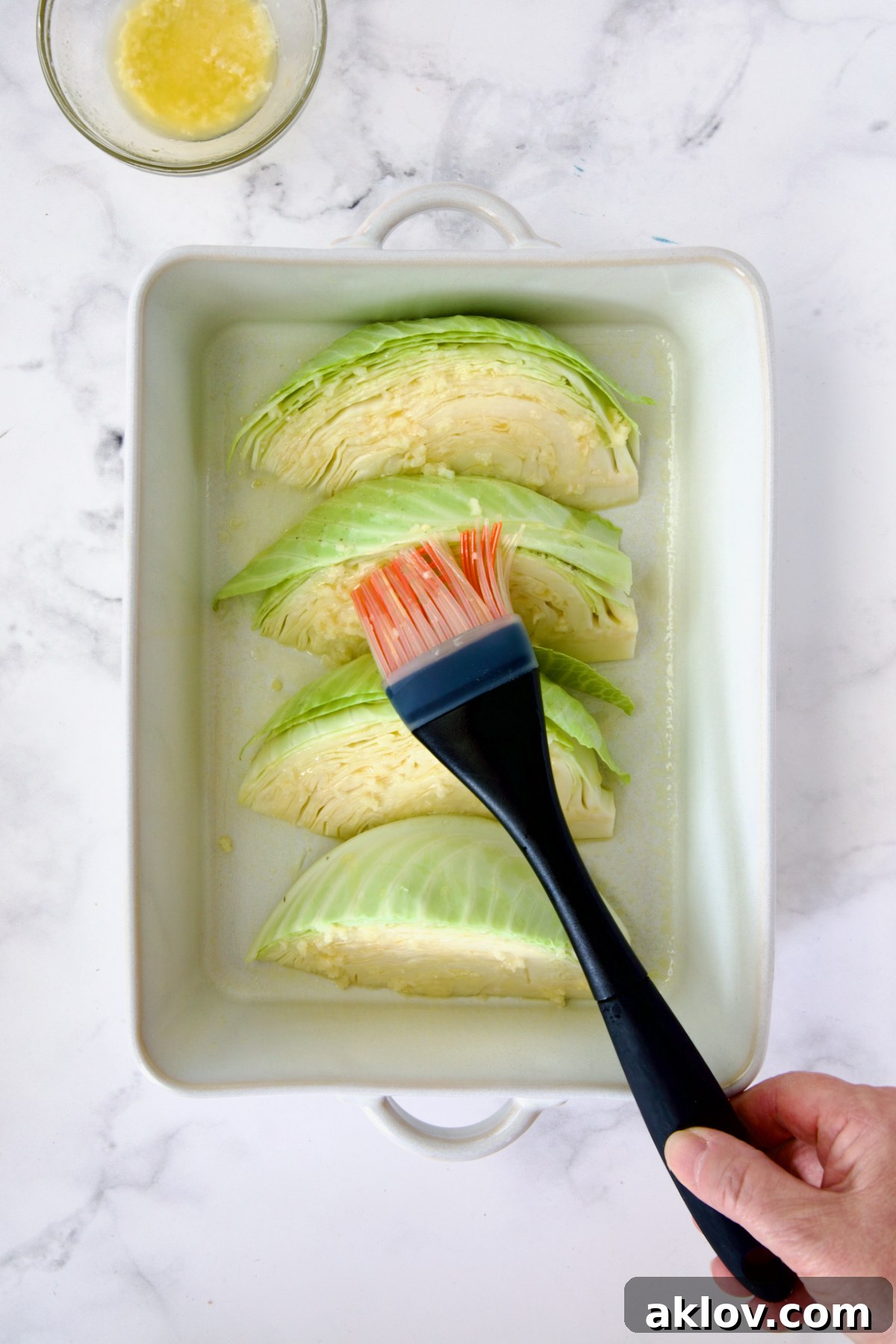 Four wedges of green cabbage in a white baking dish being brushed with melted garlic butter.
