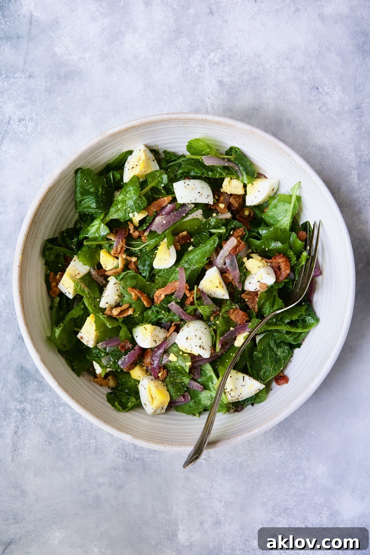 A beautifully composed Radish Greens Salad with Hot Bacon Dressing in a white serving bowl, garnished with hard-boiled eggs and a fork, ready to be enjoyed.