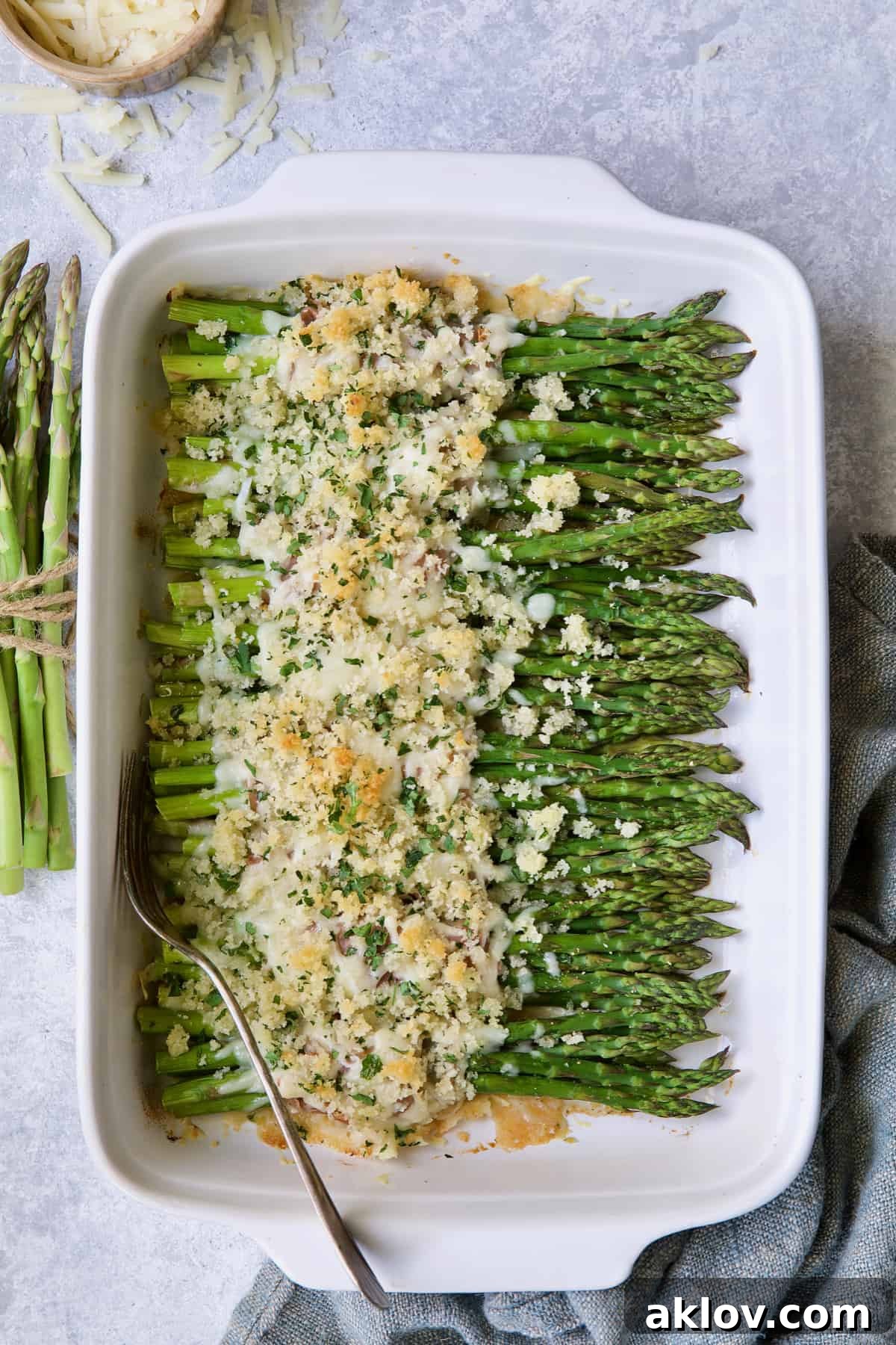 Photo of Asparagus Cordon Bleu in white rectangular baking dish with uncooked asparagus on the side and cheese scattered about.