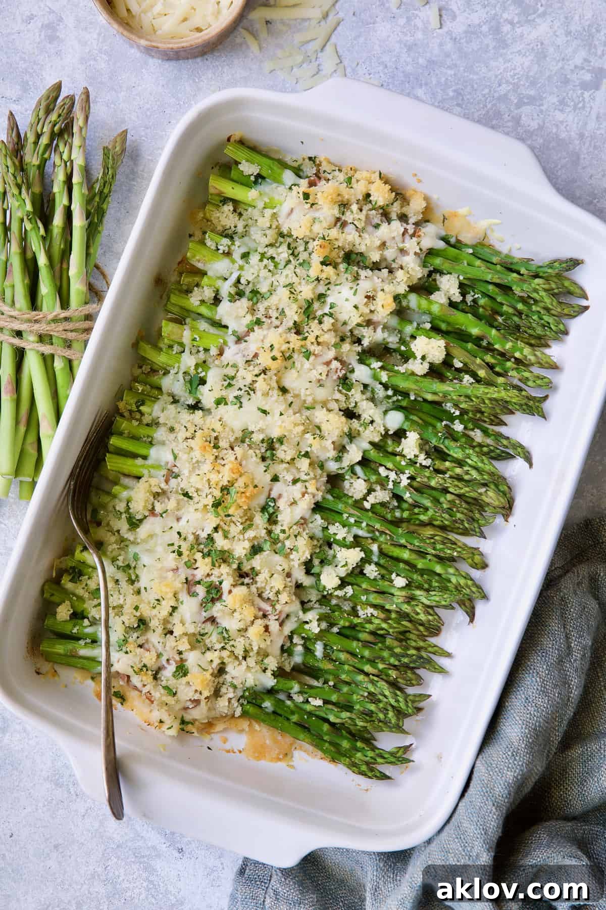 Photo of Asparagus Cordon Bleu in white baking dish on light blue background with cheese scattered about.