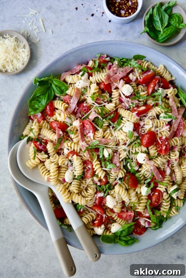 Caprese Pasta Salad artfully arranged in a blue bowl with serving utensils, flanked by small bowls of Parmesan cheese, crushed red pepper flakes, and fresh basil.