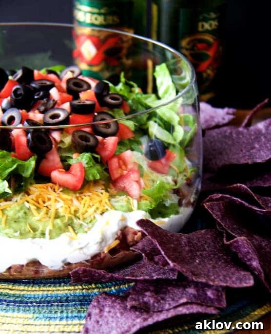 Healthier 7-Layer Dip presented in a tall glass bowl, surrounded by blue corn chips and a refreshing beer in the background.