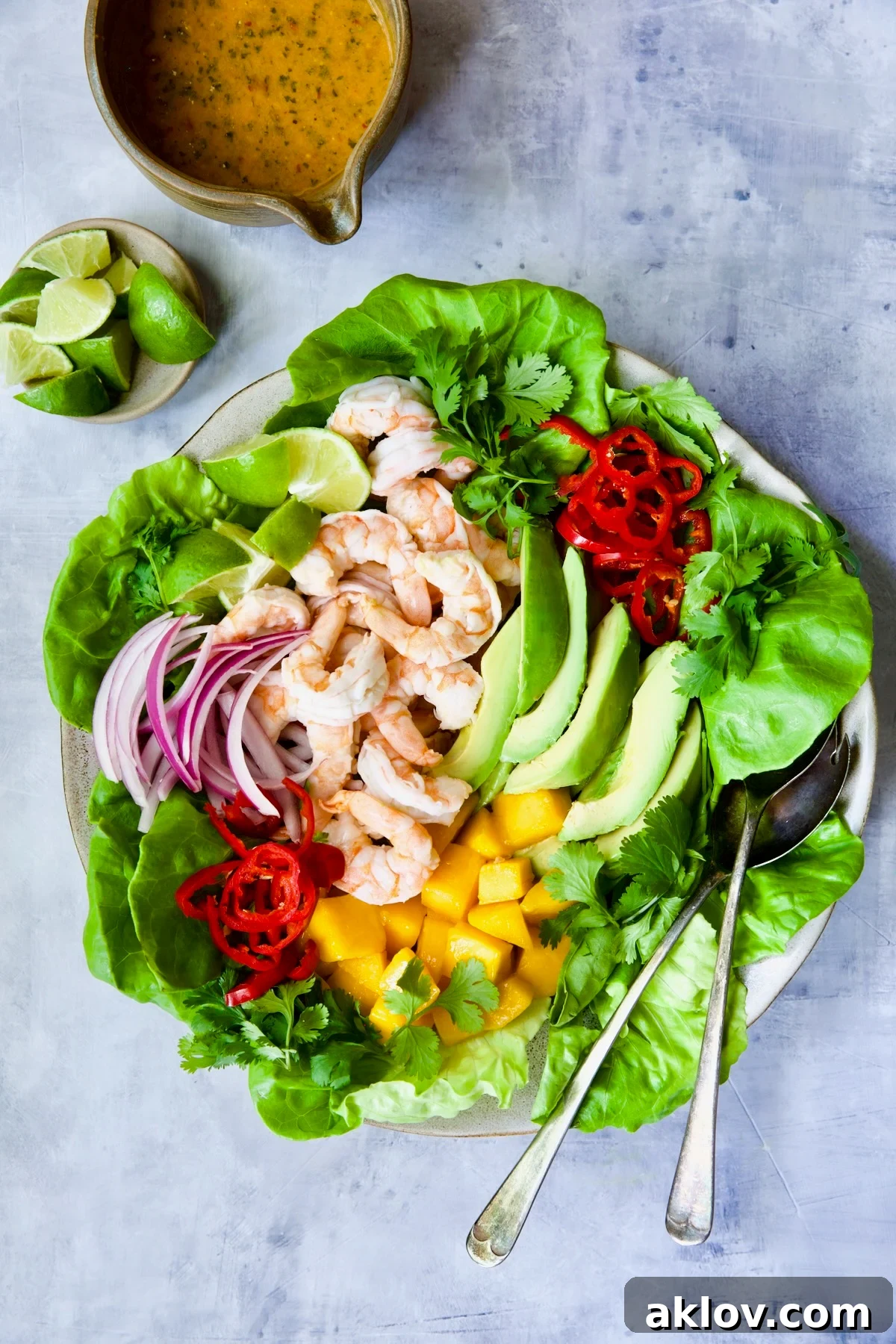 A vibrant butter lettuce salad topped with cooked shrimp, diced mango, sliced avocado, and a generous drizzle of sweet chili vinaigrette, presented on a rustic round platter with serving utensils, ready to be enjoyed as a refreshing summer meal.