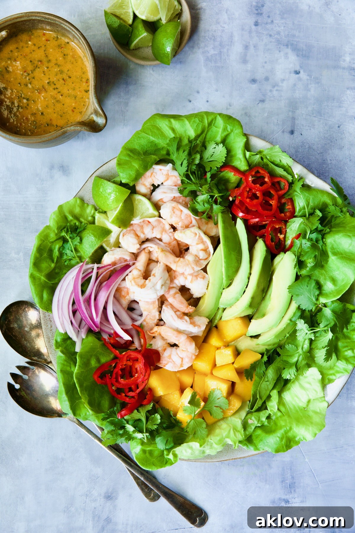 A beautifully presented Butter Lettuce Salad with Shrimp, Mango, and Avocado, garnished with lime wedges, alongside a small bowl of sweet chili vinaigrette, ready to be enjoyed as a refreshing and healthy meal.
