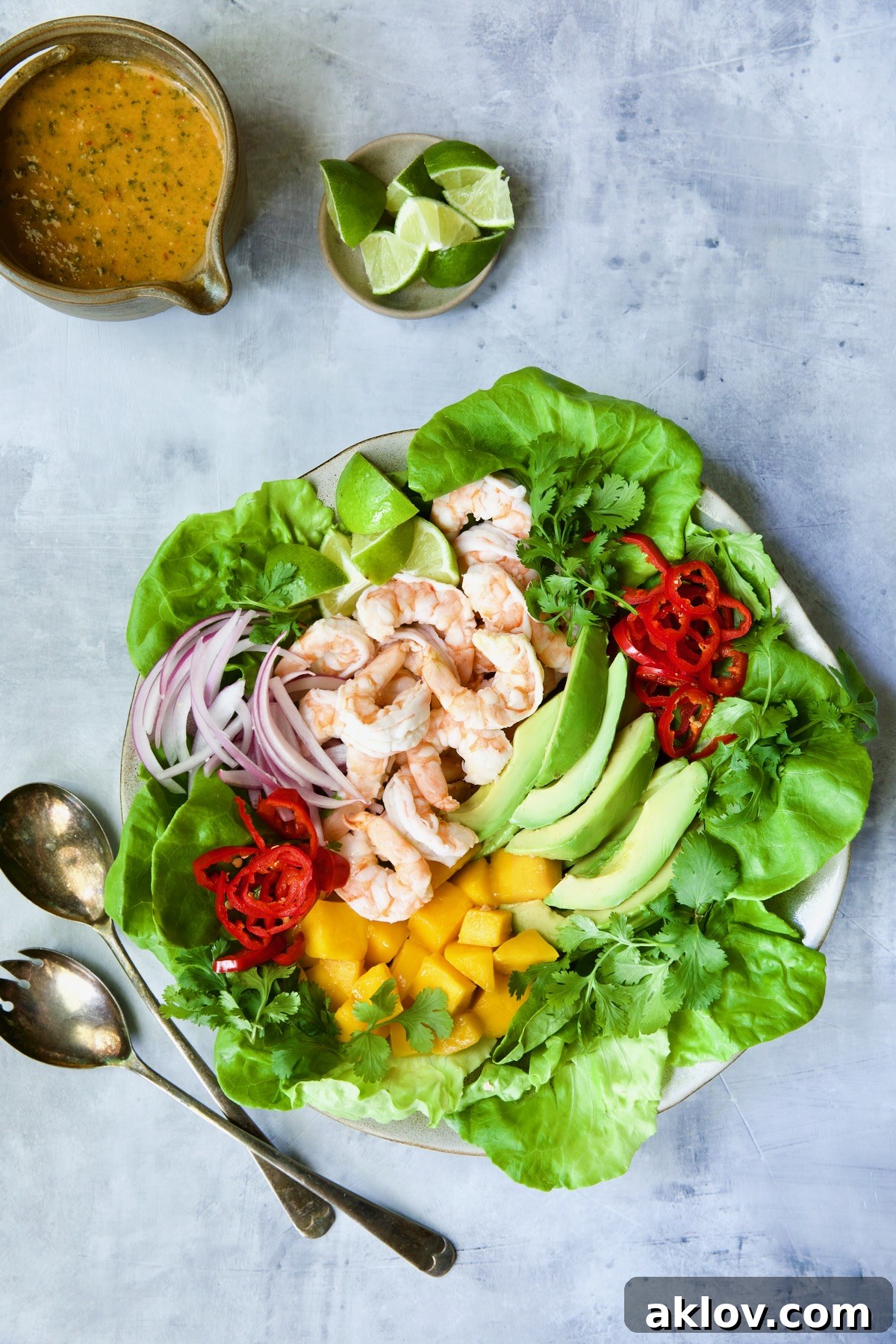 A close-up view of the butter lettuce salad, showcasing the colorful medley of shrimp, mango, and avocado, arranged artfully on a blue background, accompanied by a small bowl of sweet chili vinaigrette and fresh lime wedges.