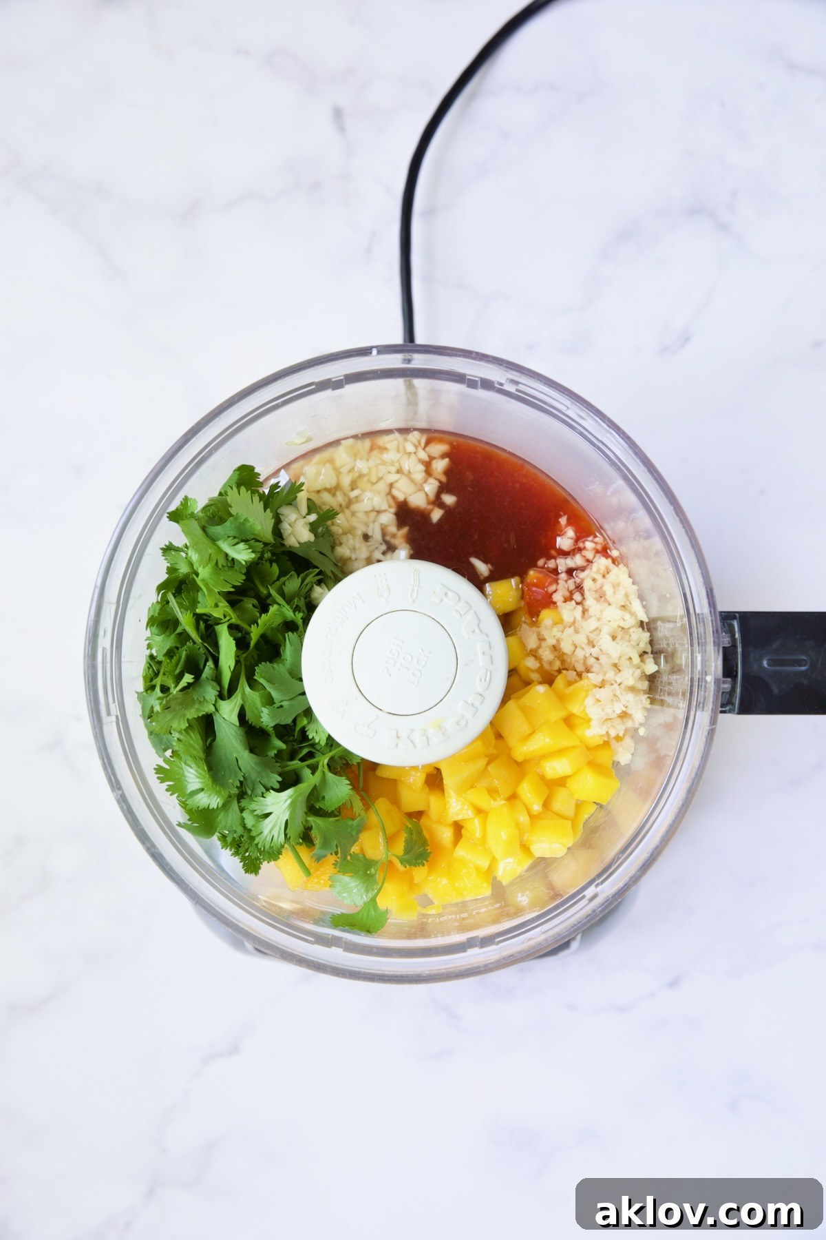 All the ingredients for the sweet chili vinaigrette, including diced mango, lime juice, ginger, garlic, cilantro, sweet chili sauce, rice vinegar, and canola oil, placed in a food processor bowl before blending.