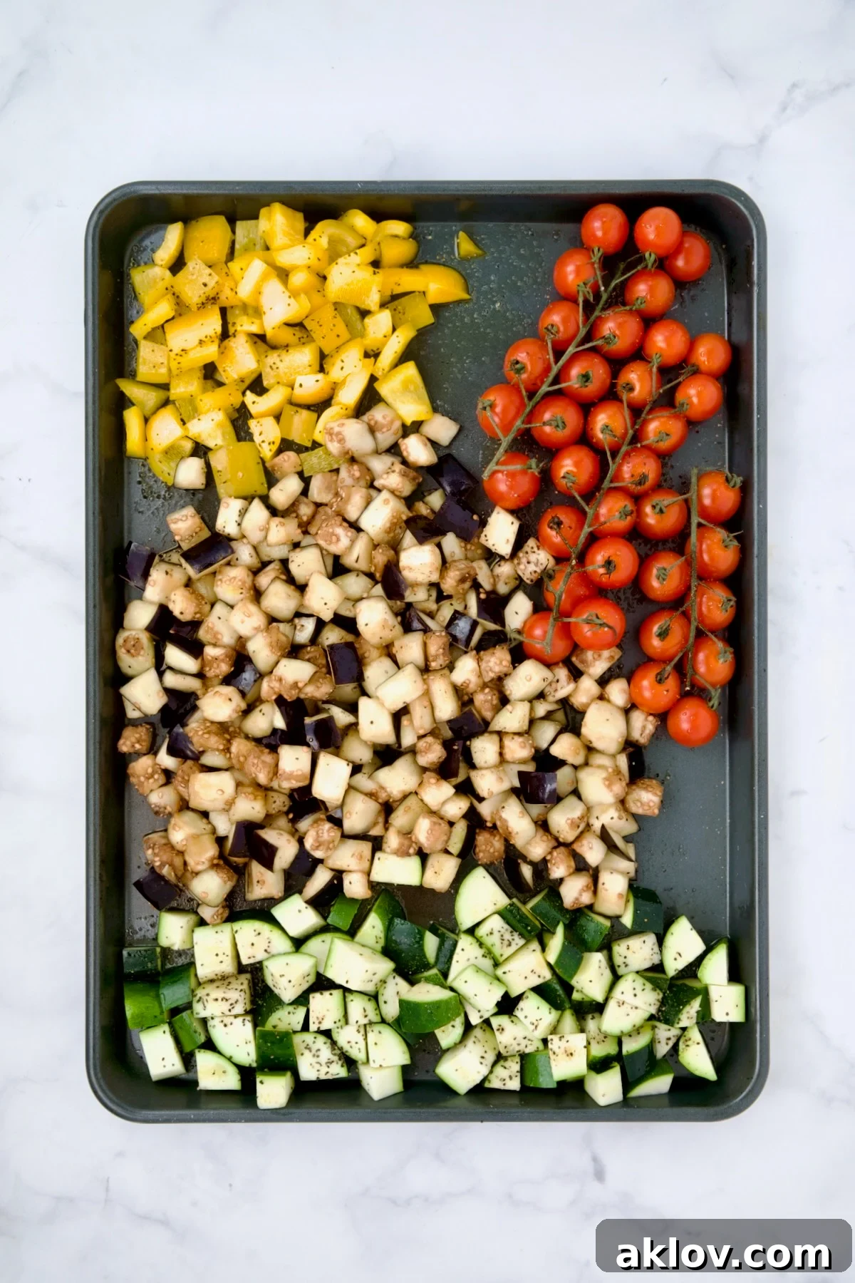 Assorted Mediterranean vegetables, including eggplant, tomatoes, zucchini, and bell pepper, arranged on a rimmed sheet pan before roasting, drizzled with olive oil.