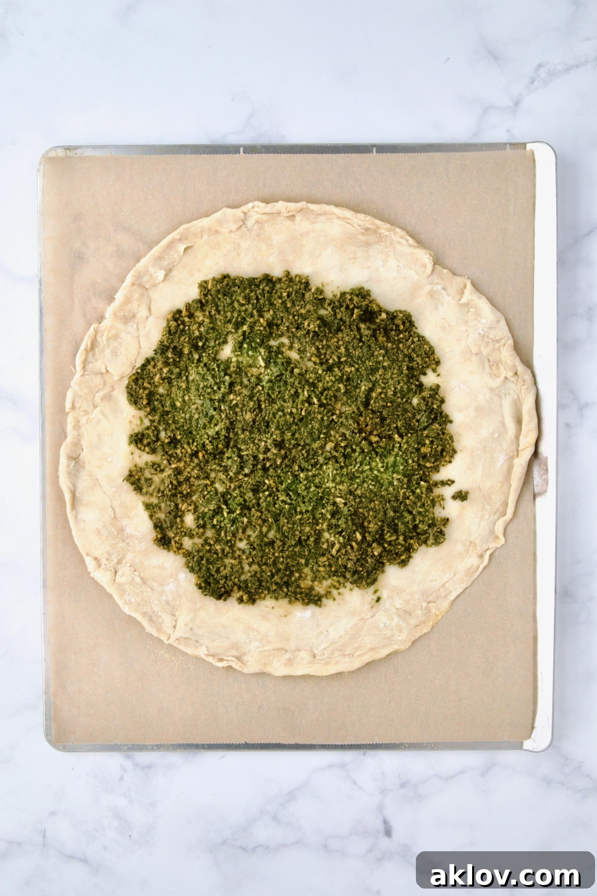 Rolled out galette dough on parchment paper on a cookie sheet, with a layer of vibrant green pesto evenly spread, leaving a border.