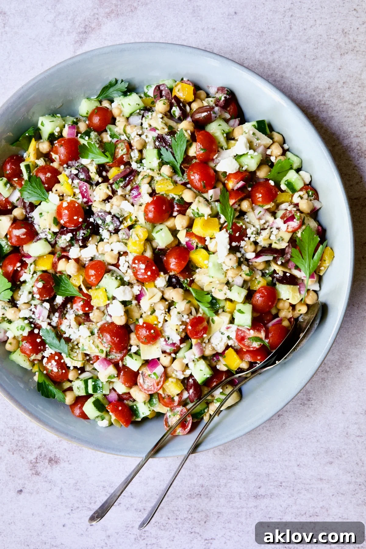 Mediterranean Chopped Salad with Chickpeas in blue bowl with serving utensils garnished with fresh parsley leaves.