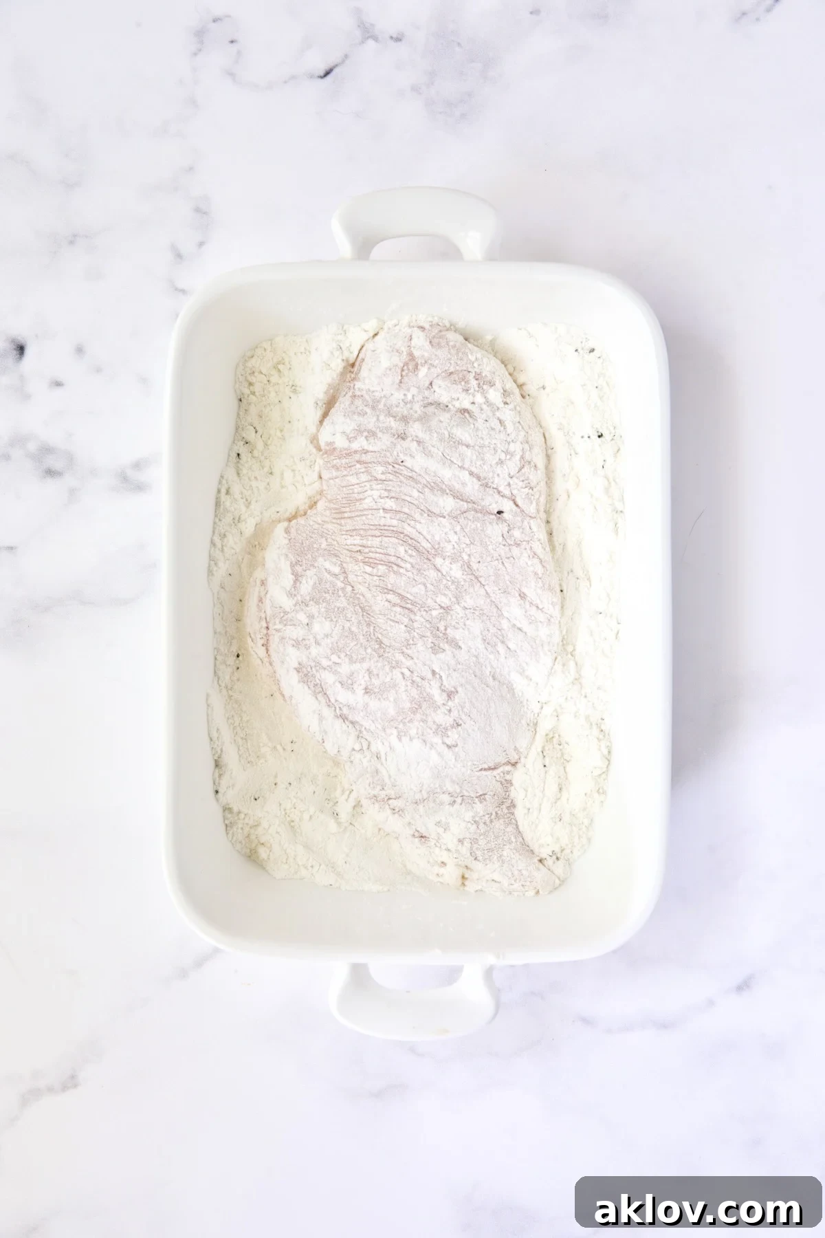 Golden Crispy Milanese 11 A raw chicken cutlet evenly coated with flour in a white dish, ready for the next breading step.