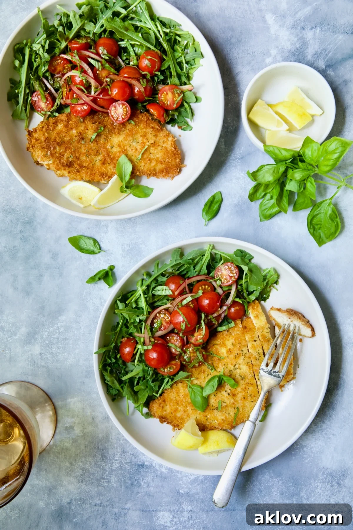 Golden Crispy Milanese 17 Two beautifully plated servings of Chicken Milanese with Arugula and Cherry Tomato Salad on white plates, garnished with fresh basil and lemon wedges.