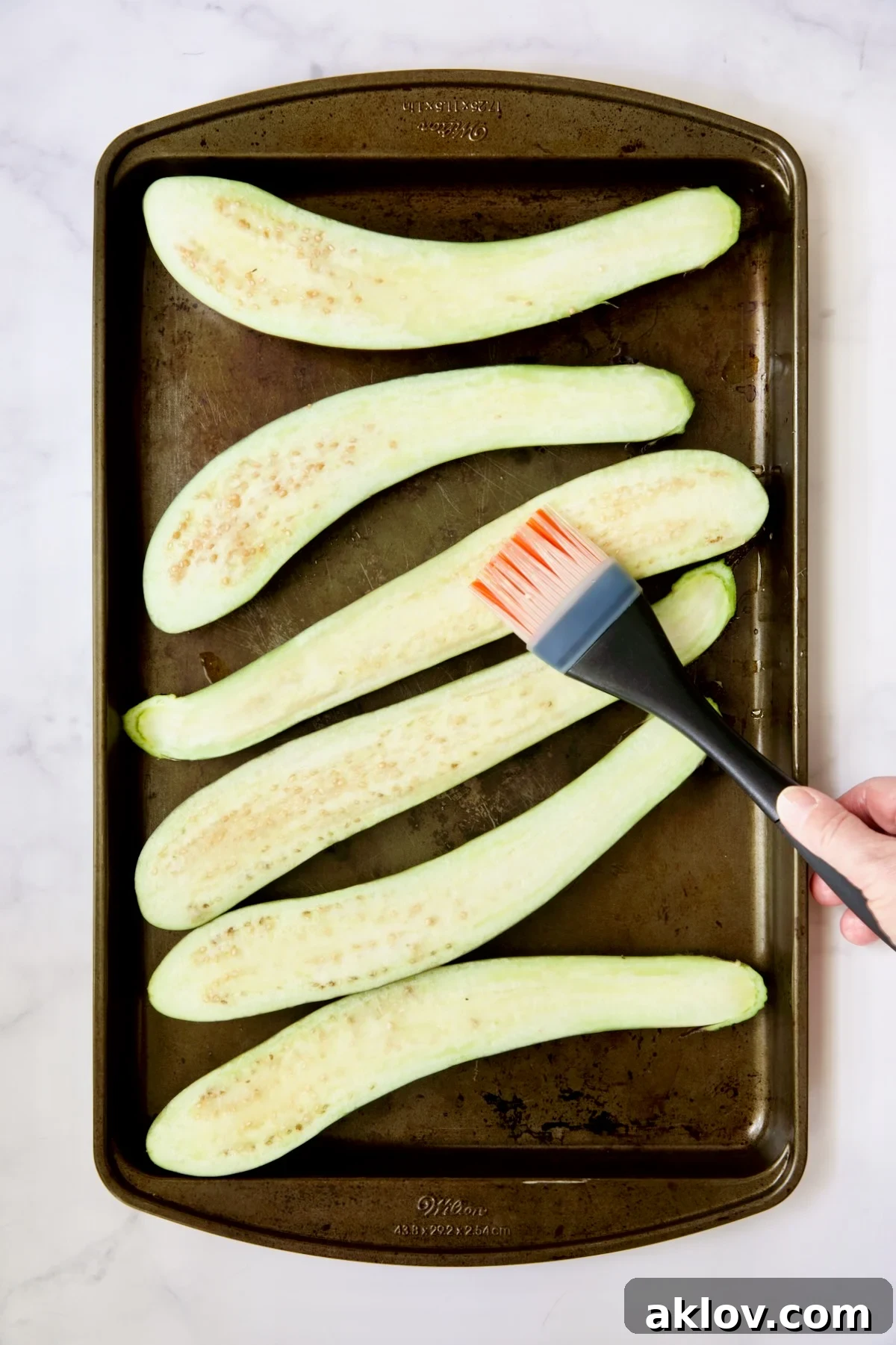 A chef brushing canola oil onto six halved Japanese eggplants arranged on a baking sheet, preparing them for roasting.