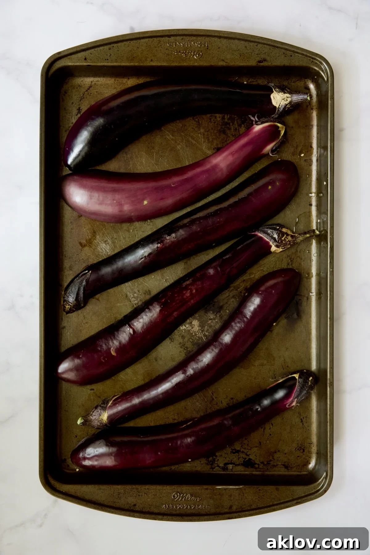 Six raw Japanese eggplant halves arranged cut-side down on a rimmed baking sheet, lightly oiled and ready for baking.