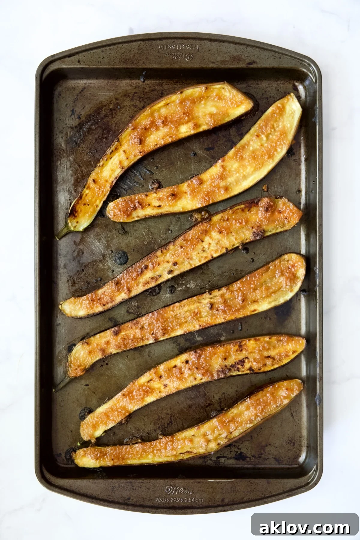 Six perfectly broiled Japanese eggplant halves with a glossy, caramelized miso glaze, resting on a stainless steel rimmed baking sheet, ready for garnishing.