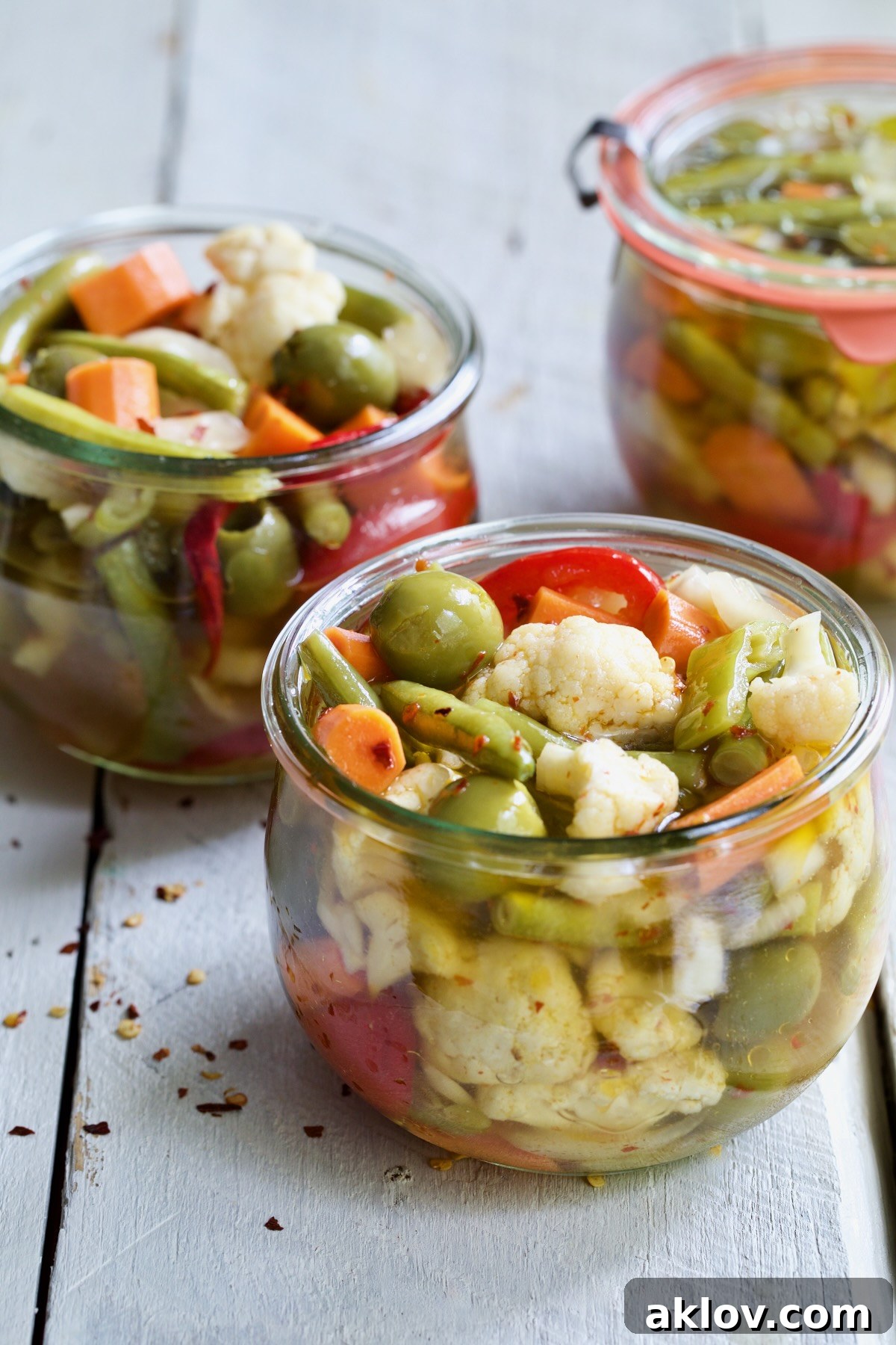 Three glass jars of freshly made Giardiniera (Italian Pickled Vegetables) lined up on a rustic white wooden surface, showcasing their appealing colors and textures.