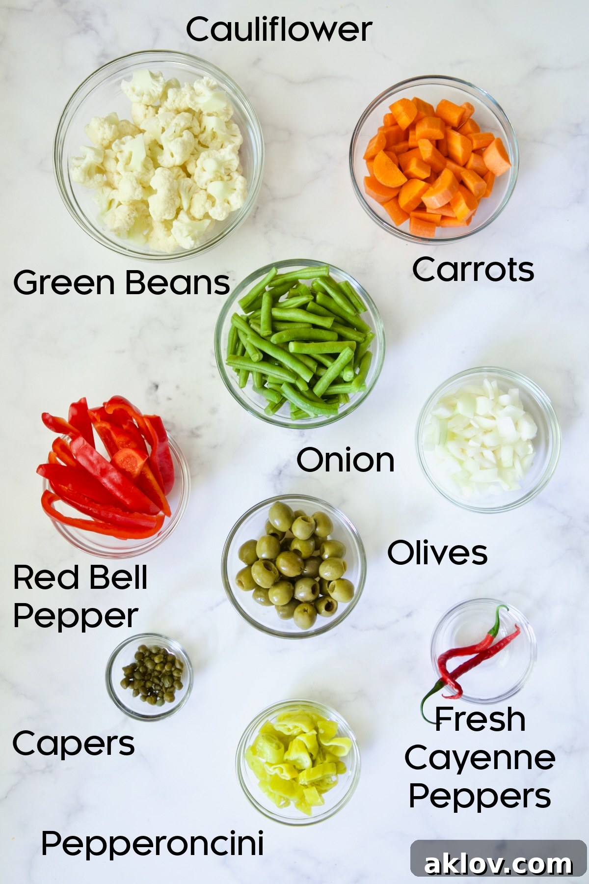 Separate glass bowls displaying the fresh vegetables for Giardiniera: cauliflower florets, sliced carrots, trimmed green beans, red bell pepper strips, chopped onion, green olives, capers, and cayenne peppers.