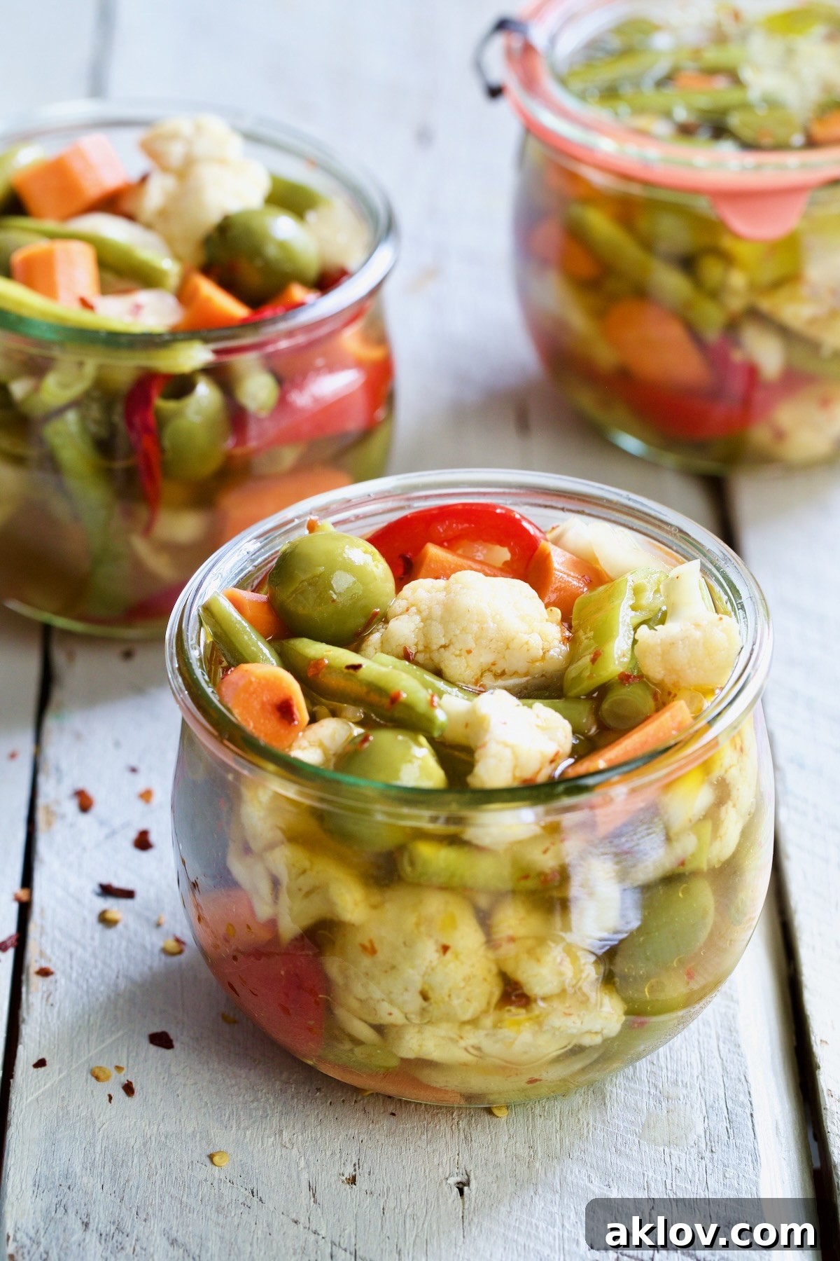 Three perfectly prepared jars of Giardiniera (Italian Pickled Vegetables) on a white wooden table, showcasing the final product.