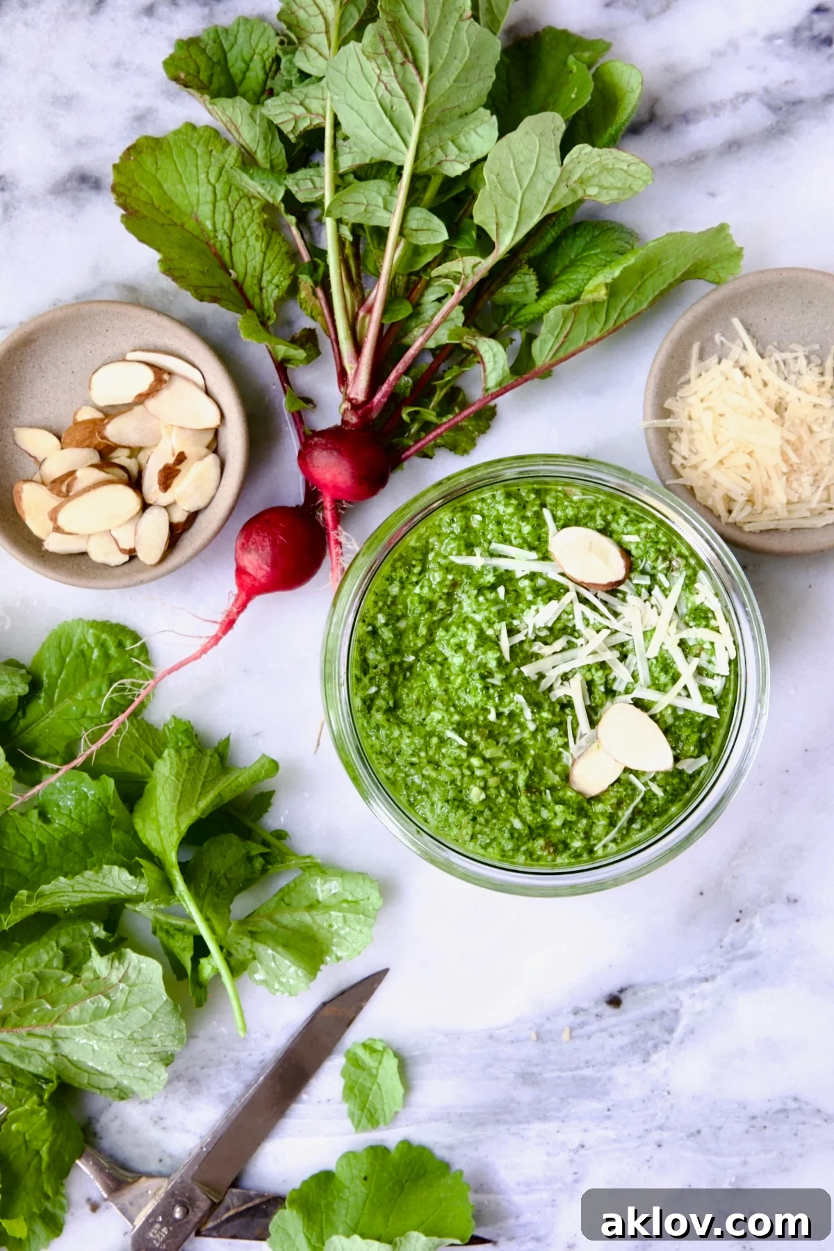 Close-up of Radish Greens Pesto in a glass jar with a small spoon, set on a marble surface. Ingredients like whole almonds, garlic cloves, and fresh radish greens are artfully scattered around, creating an inviting culinary scene.
