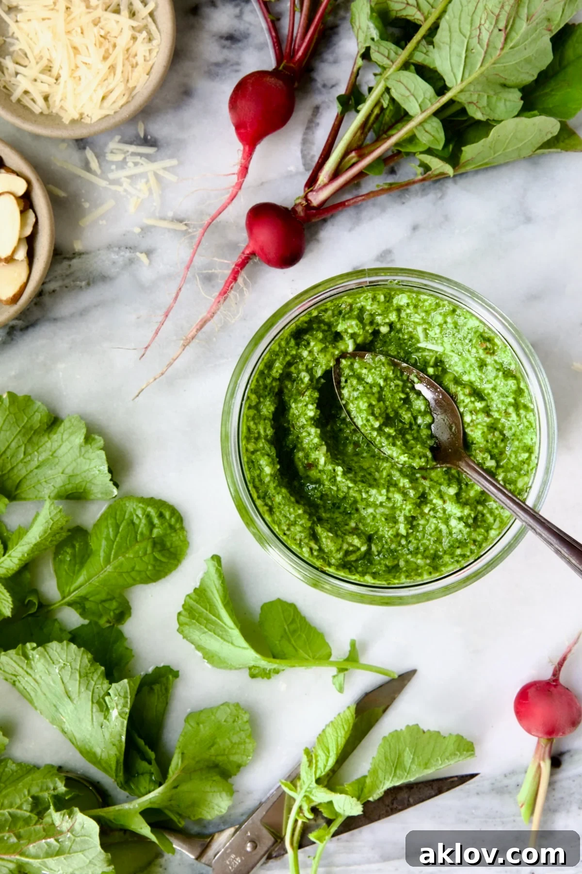 Radish Greens Pesto in glass jar with spoon on marble surface with ingredients in the pesto around it.