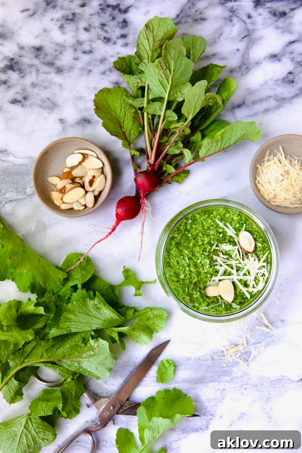 Radish Greens Pesto in glass jar with spoon on marble surface with ingredients in the pesto around it.