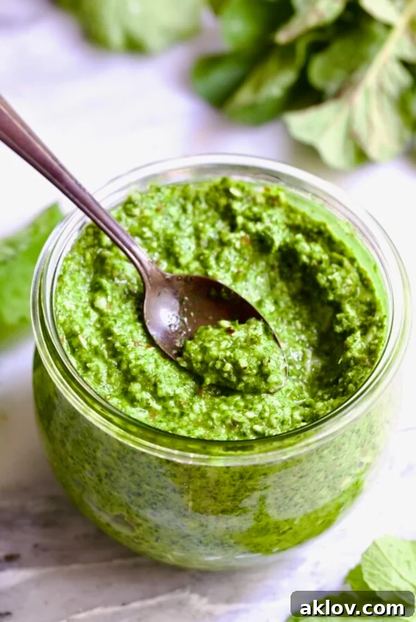A close-up shot of vibrant green Radish Greens Pesto in a glass jar, with a small spoon scooping out a generous portion, emphasizing its rich texture and appealing color.