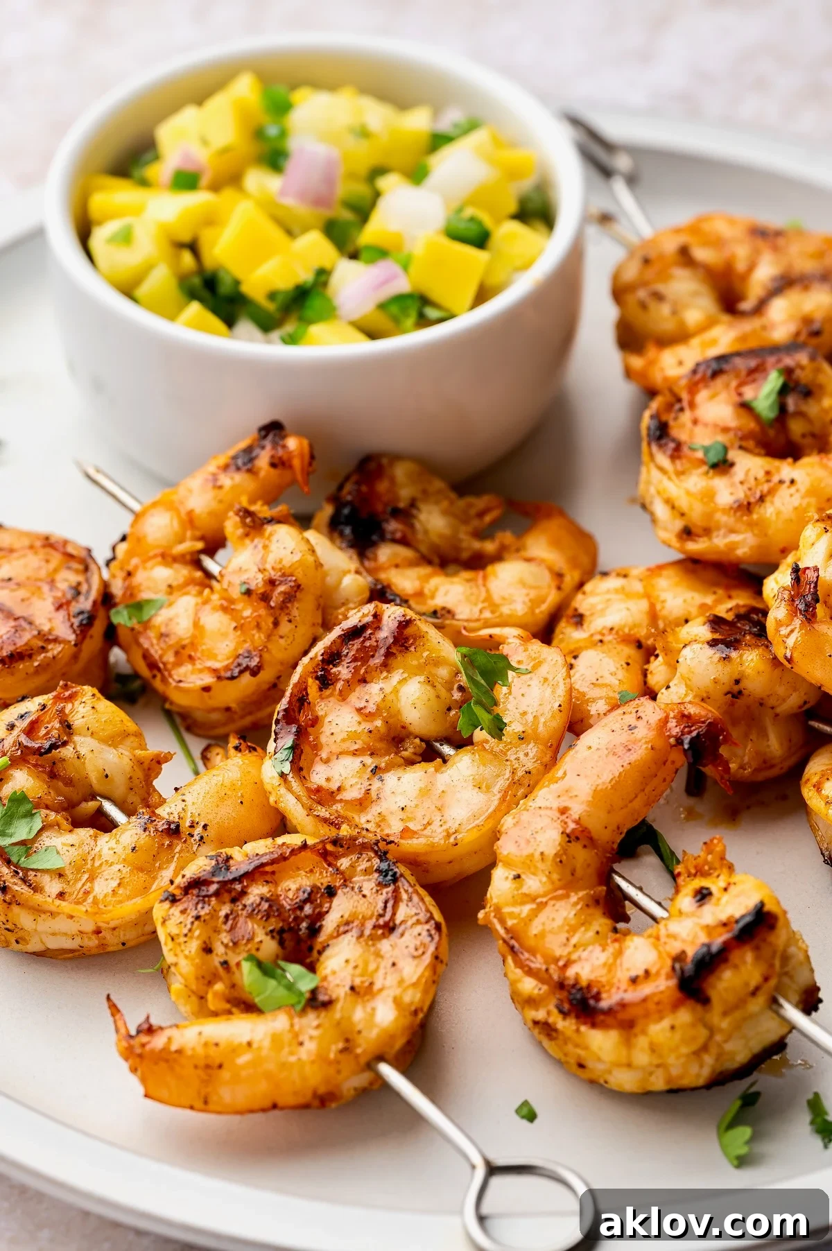 A beautifully presented dish featuring grilled shrimp skewers on a white plate, accompanied by mango pineapple salsa in a small white bowl.