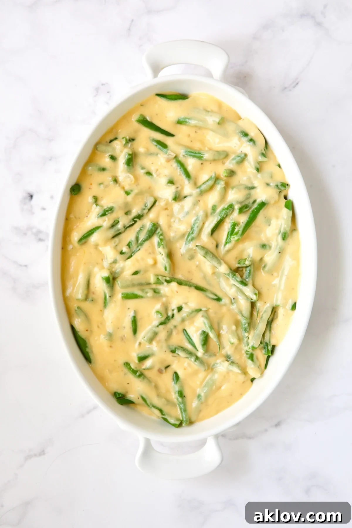 Fresh green beans generously covered with a rich, creamy cheese sauce in a white oval gratin dish, pre-baking.