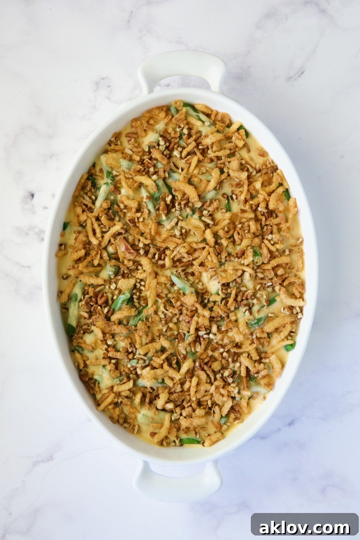Irresistible Cheesy Green Bean Bake 12 Unbaked cheesy green bean casserole in a gratin dish, fully assembled with pecans and fried onions on top.