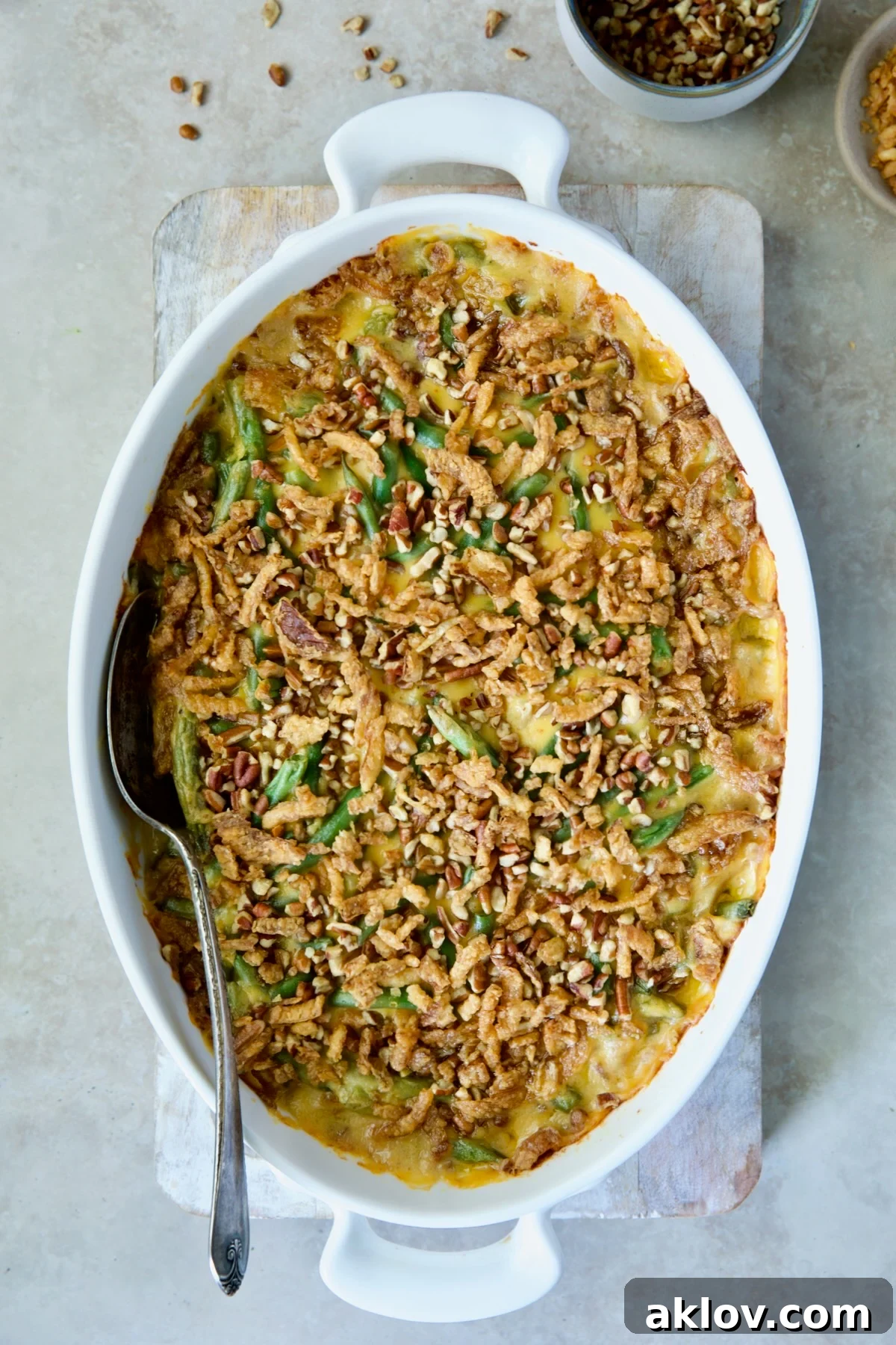 A freshly baked Cheesy Green Bean Casserole, glistening with a golden-brown pecan and crispy onion topping, presented on a wooden cutting board with a serving spoon.