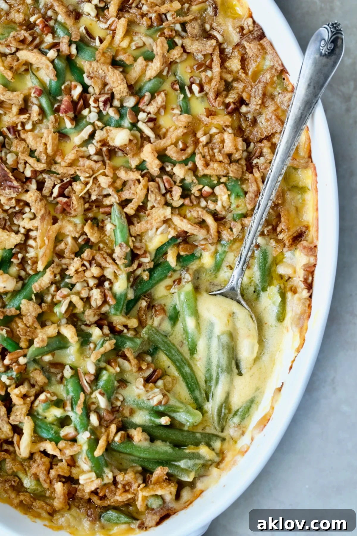 A freshly baked Cheesy Green Bean Casserole, showcasing its rich golden topping and creamy interior, resting in a white oval gratin dish with a serving spoon nestled beside it.