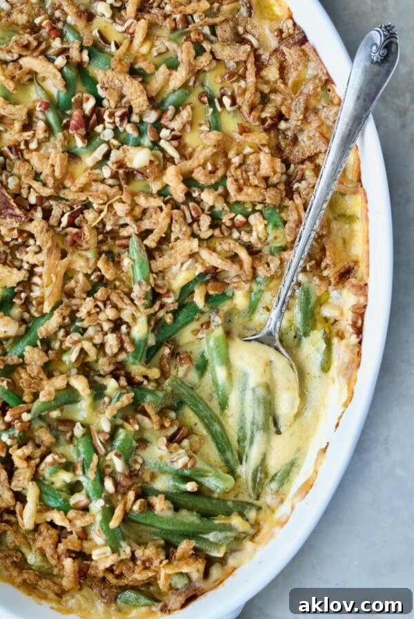Cheesy Green Bean Casserole in a white oval gratin dish with a serving spoon, showcasing its rich topping and creamy interior.
