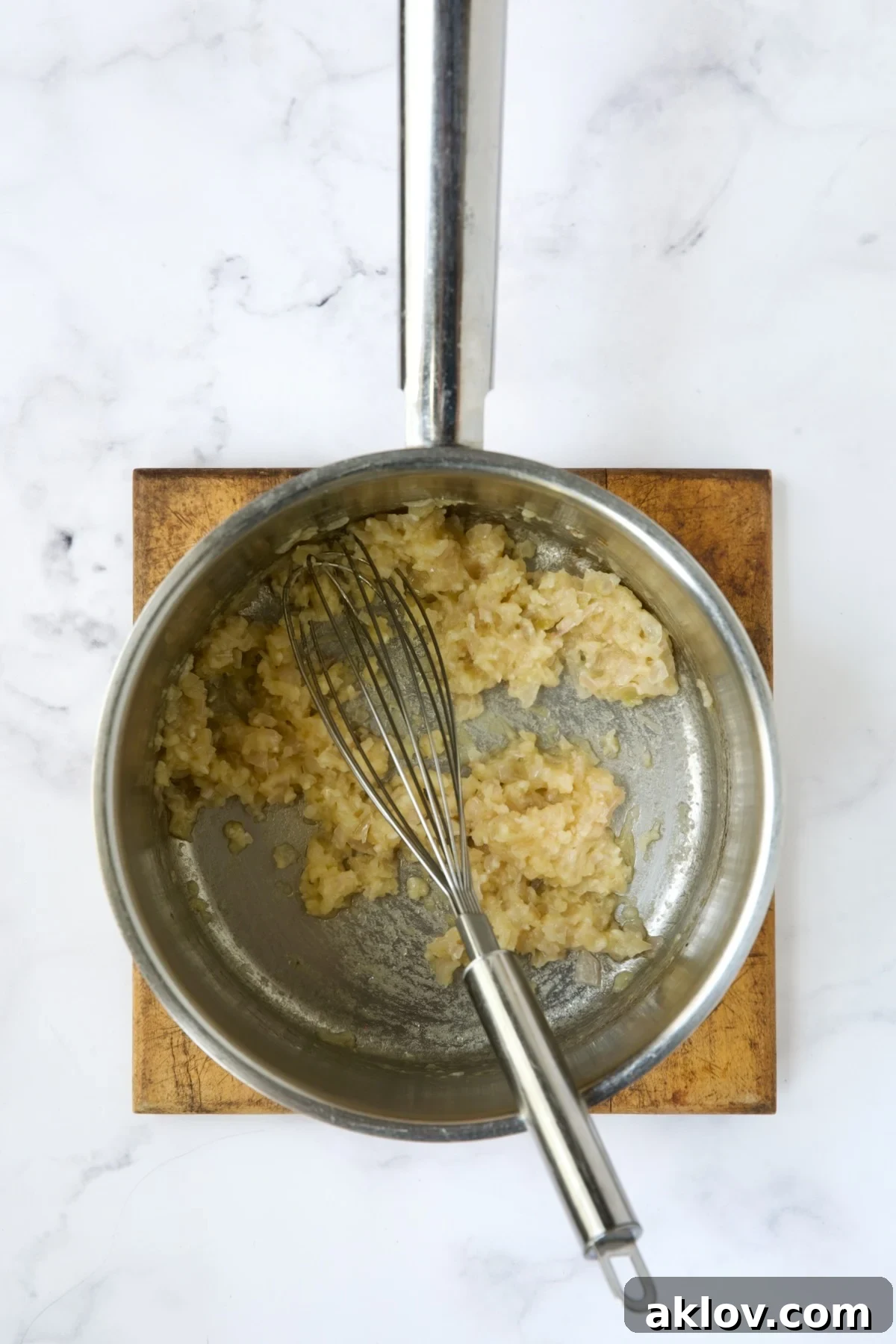 A wire whisk expertly blending shallots, garlic, and flour in a stainless steel saucepan, forming the base for a rich, creamy sauce.