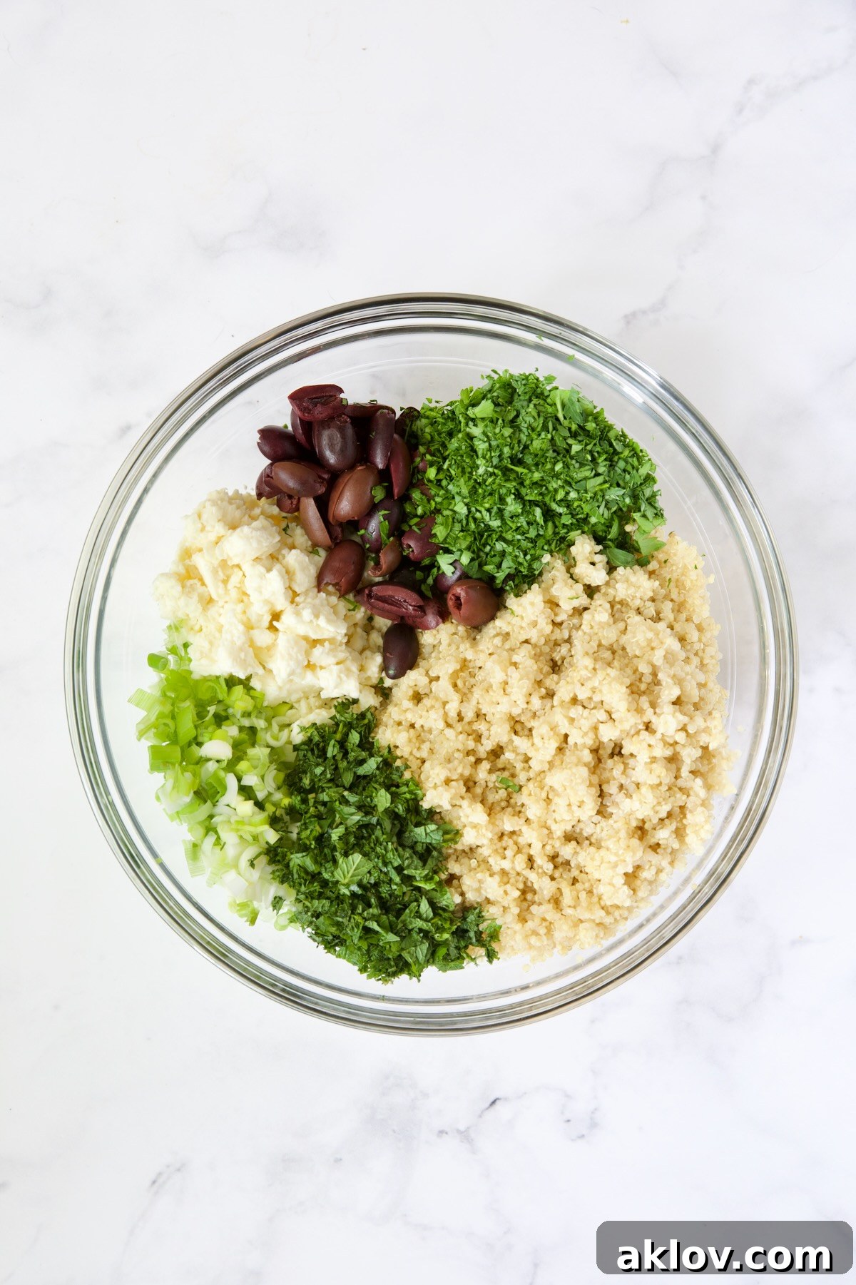 A colorful medley of cooled quinoa, olives, feta cheese, scallions, fresh mint, and parsley mixed together in a large glass bowl.