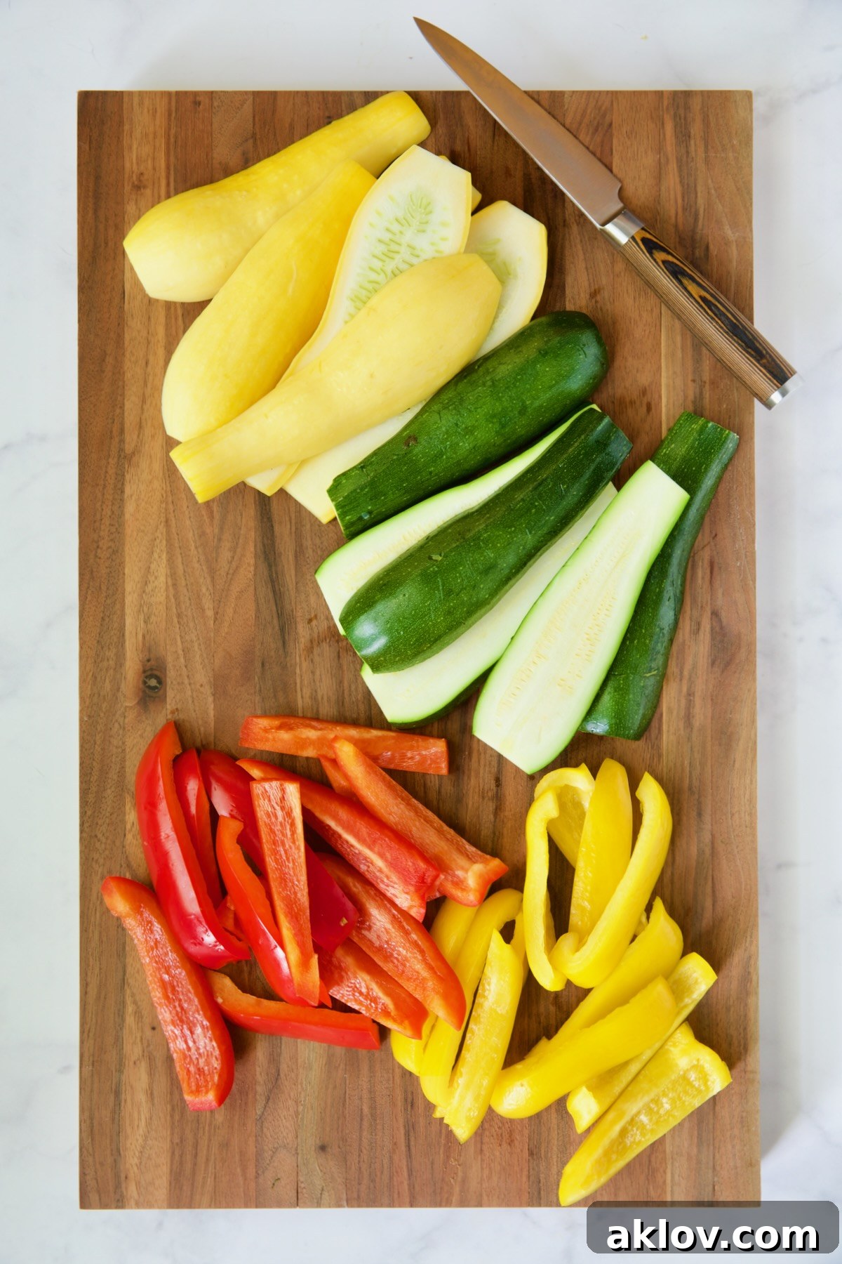 Freshly cut zucchini, yellow squash, and bell peppers neatly arranged on a wooden cutting board, alongside a sharp knife, prepared for grilling.