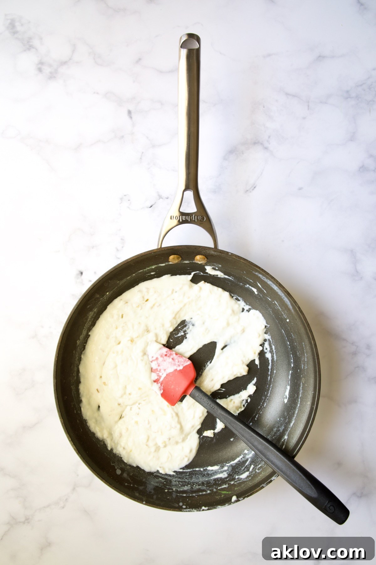 Thickened half-and-half sauce being stirred in a nonstick skillet with a red and black rubber spatula.