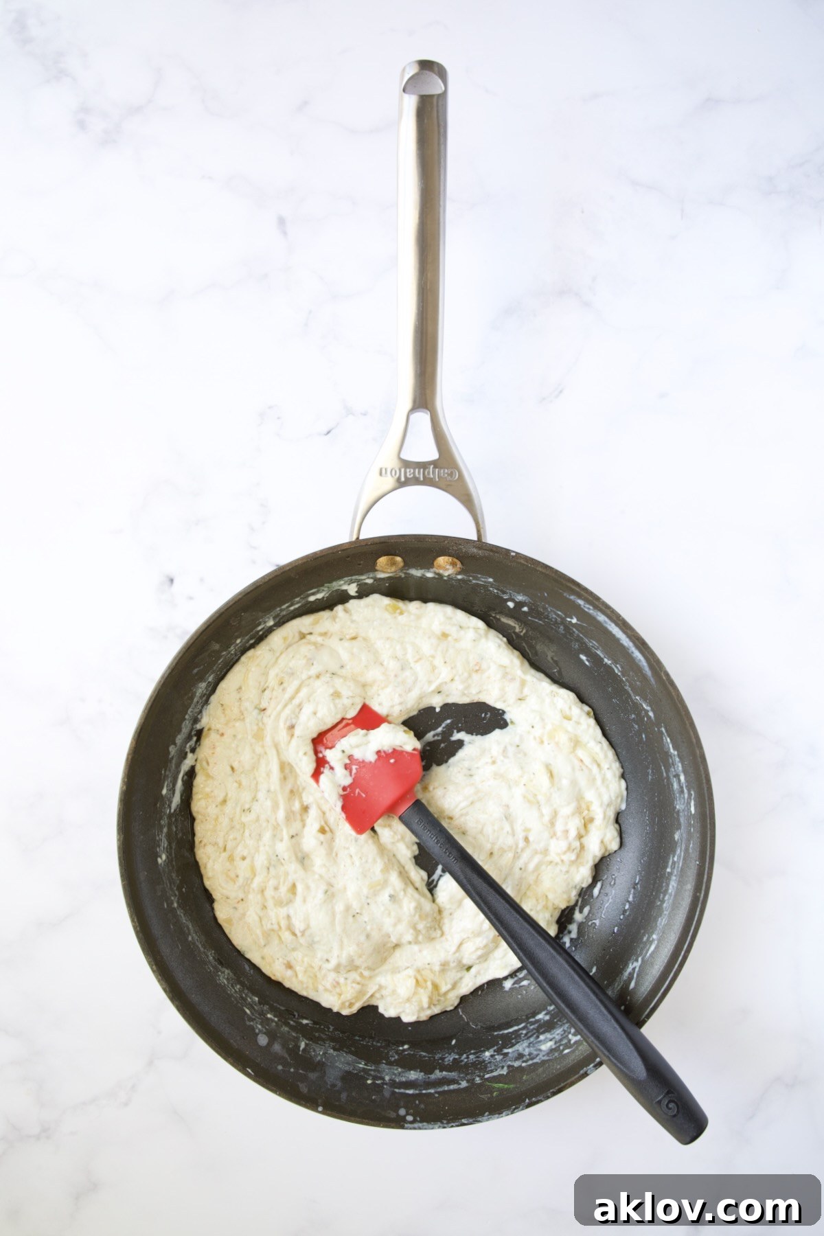 Thickened cheese sauce in a nonstick skillet being stirred with a red and black spatula.