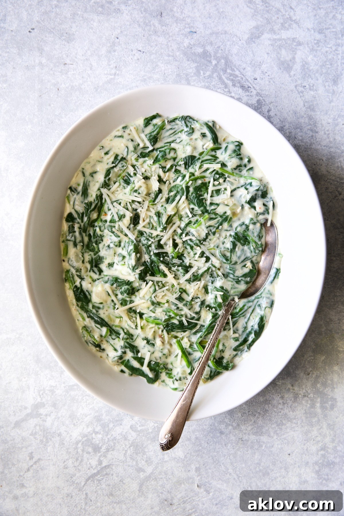 A bowl of Steakhouse Creamed Spinach with a serving spoon on a light blue-gray background.