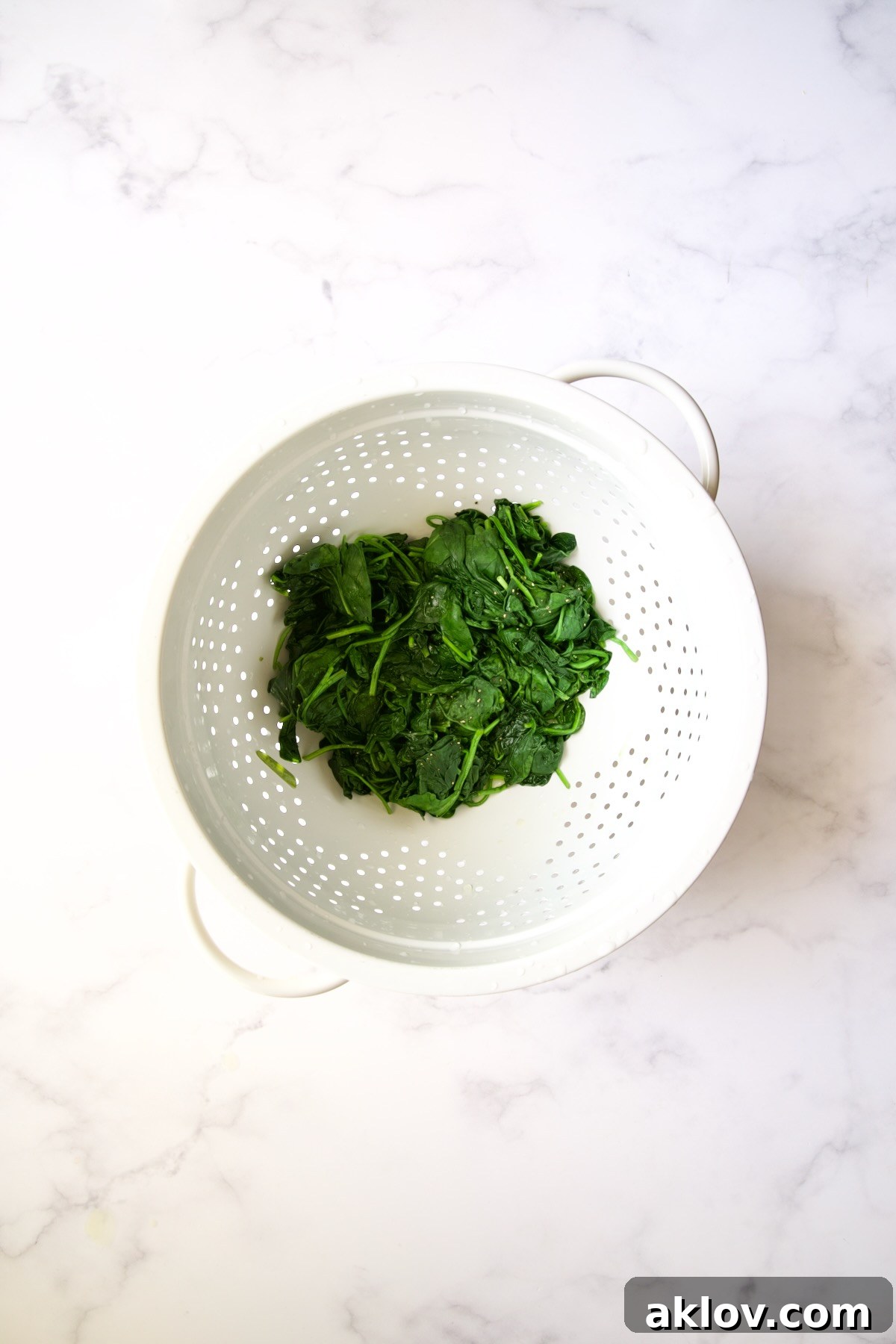 Wilted baby spinach being pressed in a white colander to remove excess water.