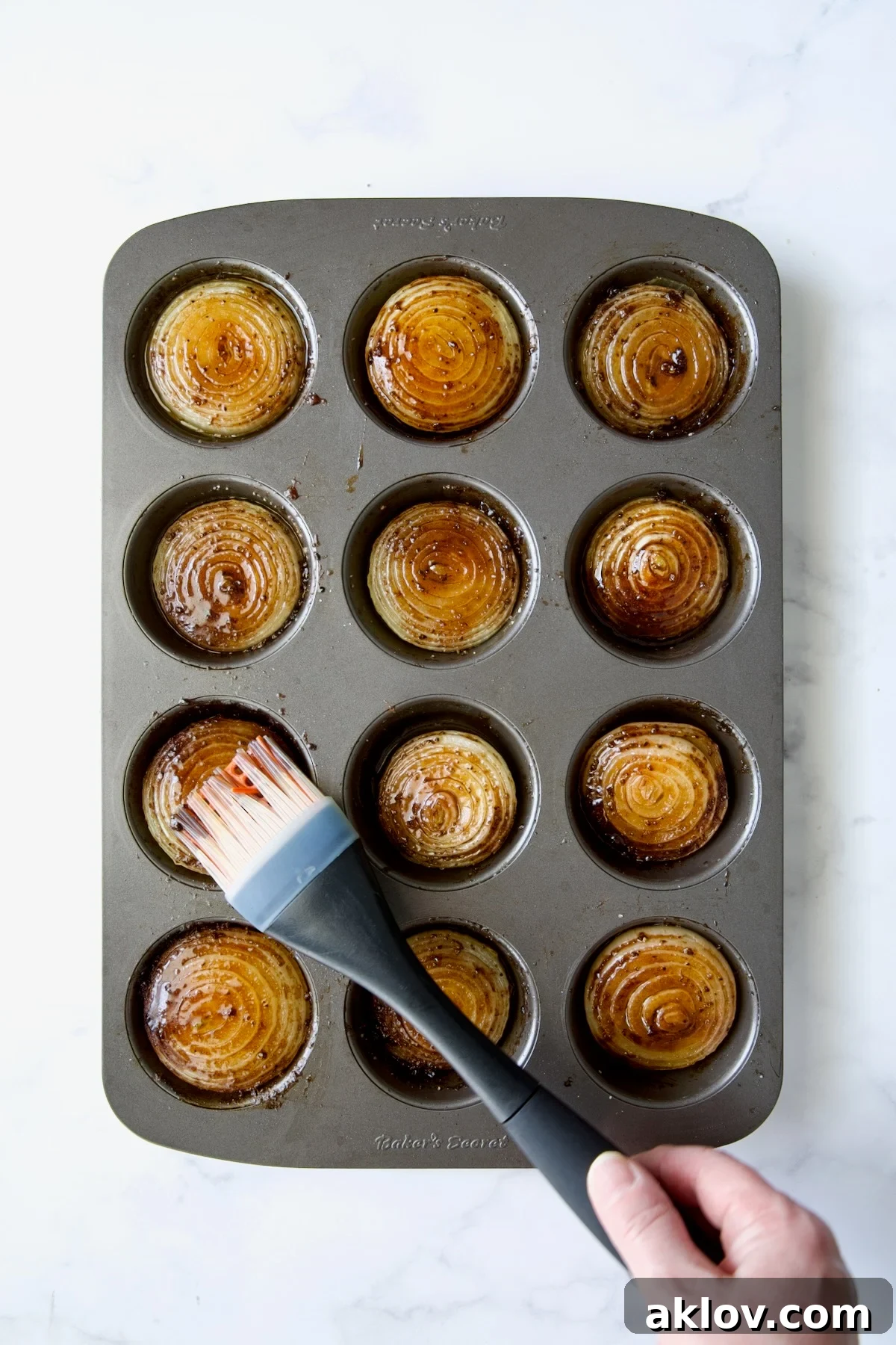 A pastry brush smoothing the mixture of ghee and beef base over the onion halves in a nonstick muffin pan, ensuring even coating.