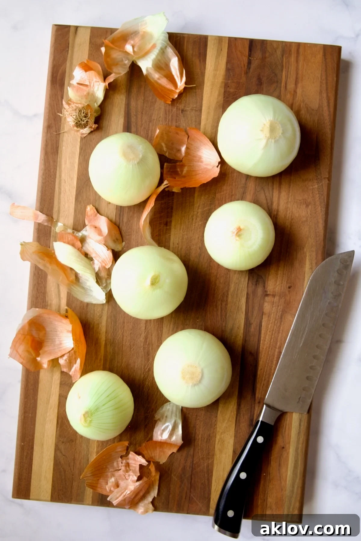 Six peeled onions with some of the discarded peels and a chef's knife resting on a wooden cutting board, ready for preparation.