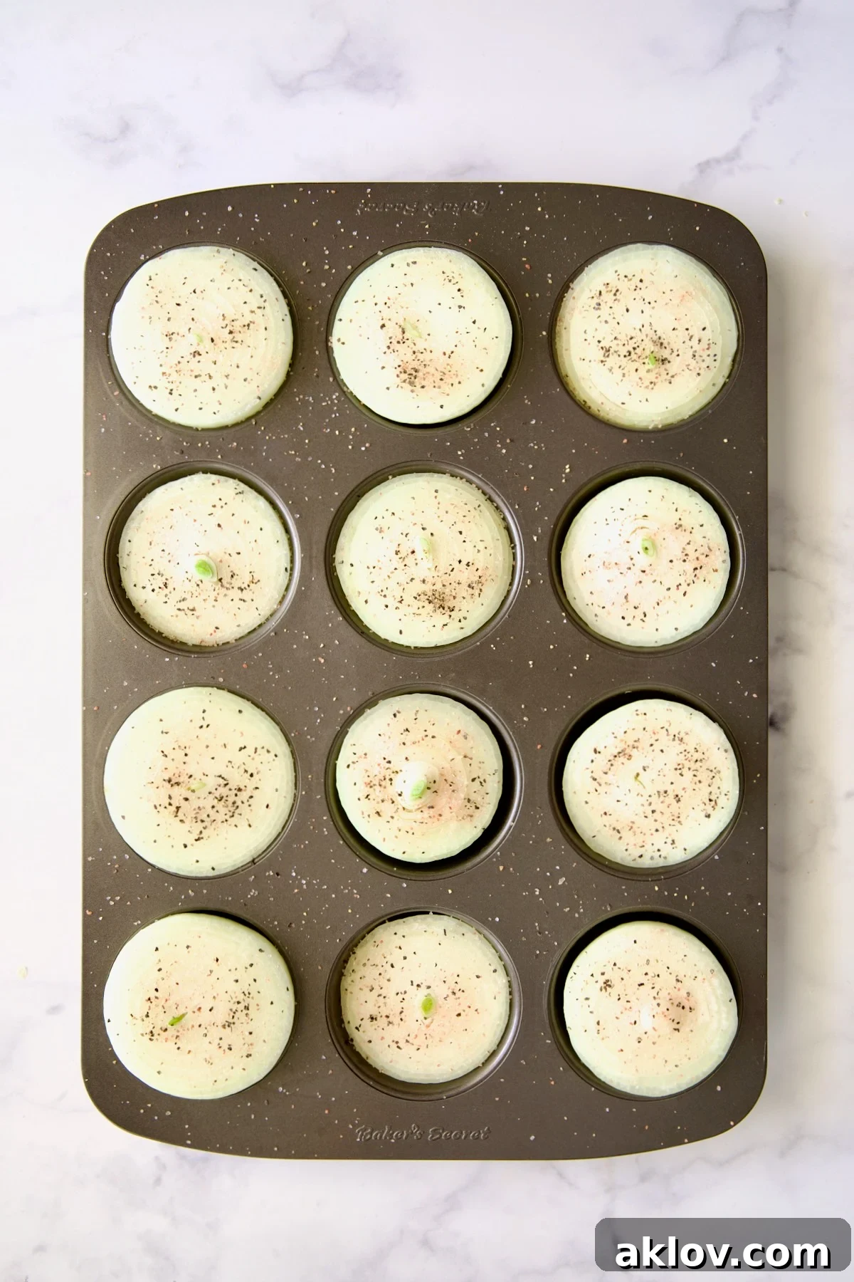 Onion halves neatly arranged in a nonstick muffin pan, with their cut sides facing up, lightly seasoned with black pepper.