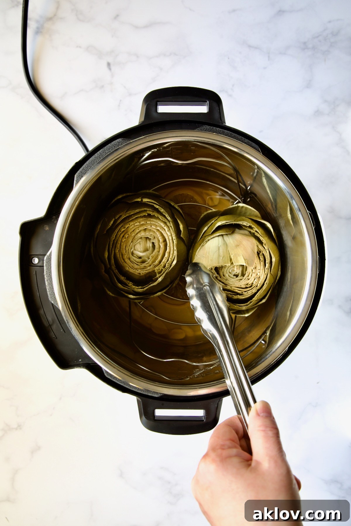 A pair of tongs gently pulling a leaf from one of the two steamed artichokes inside the Instant Pot, demonstrating doneness.