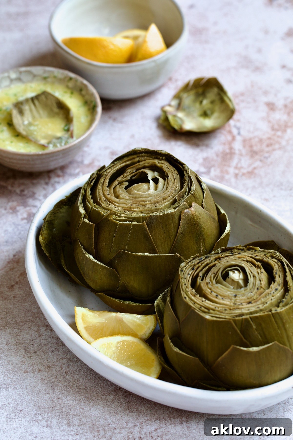 Artichokes prepared in the Instant Pot, presented in an elegant white oval serving bowl, with a small dish of garlic butter and lemon wedges blurred softly in the background.