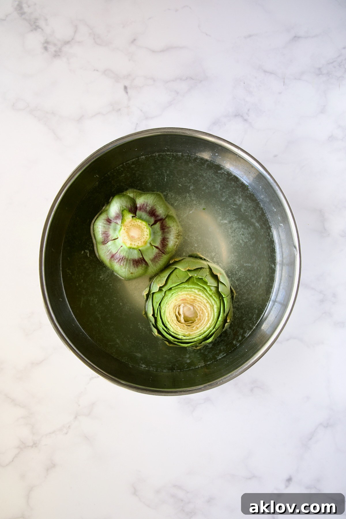Prepared artichokes submerged in a stainless steel bowl with lemon water, preventing oxidation.