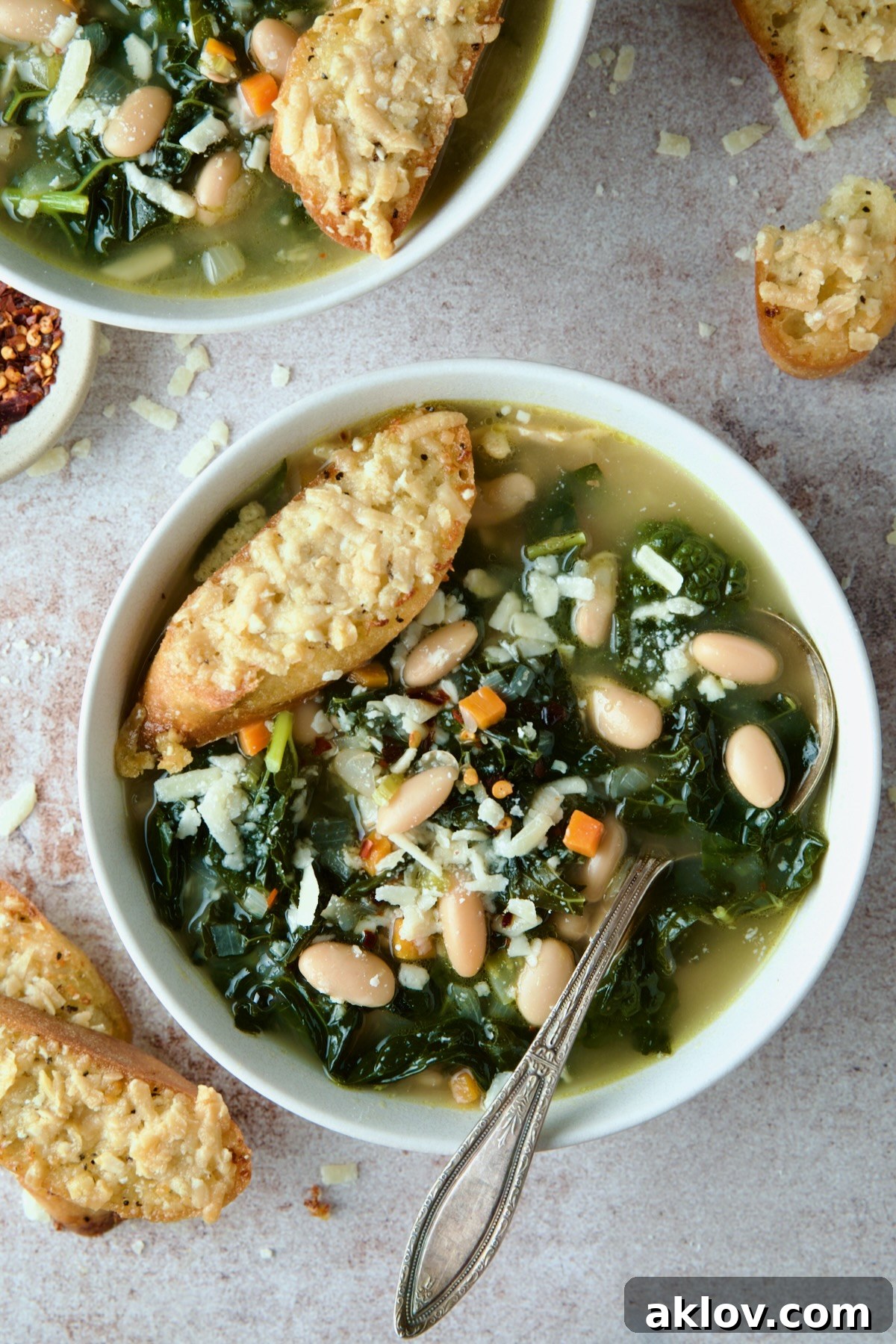 A steaming bowl of rustic White Bean Kale Soup, garnished with a sprinkle of fresh herbs, accompanied by a golden-brown Parmesan Toast, resting invitingly on a white surface.