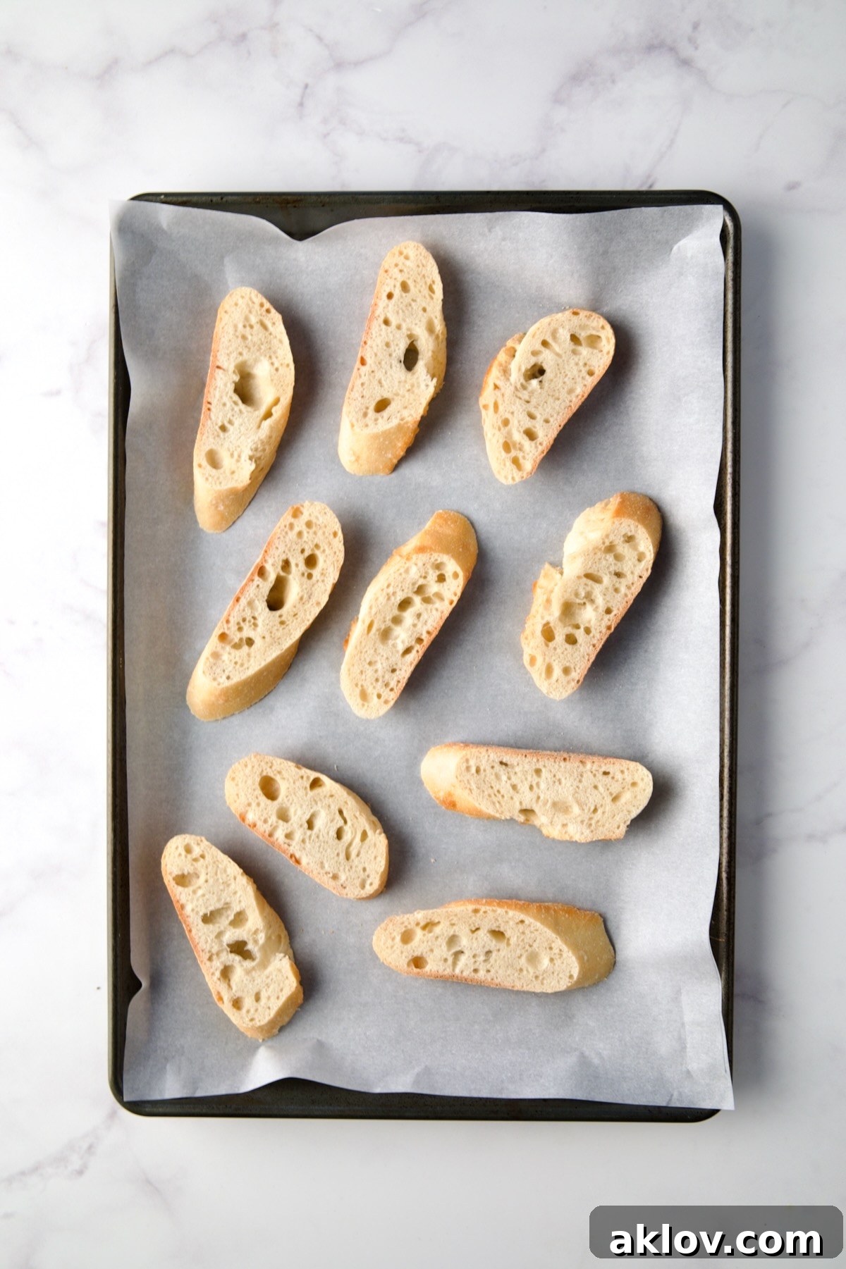 Slices of baguette are neatly arranged on a parchment paper-lined rimmed baking sheet, prepared for the Parmesan Toast topping.
