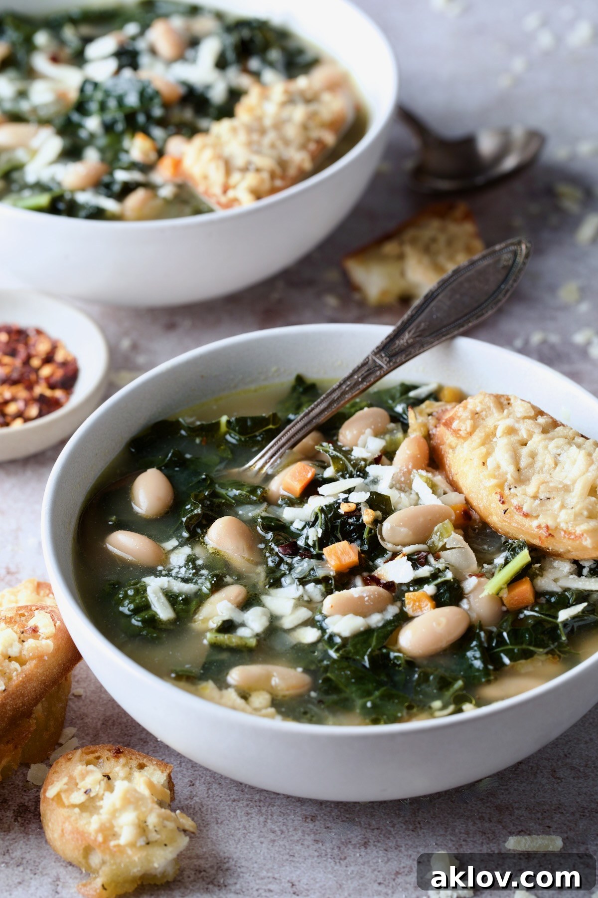 A beautifully presented bowl of White Bean Kale Soup, with Parmesan Toast perfectly balanced on top, another bowl blurred in the background.