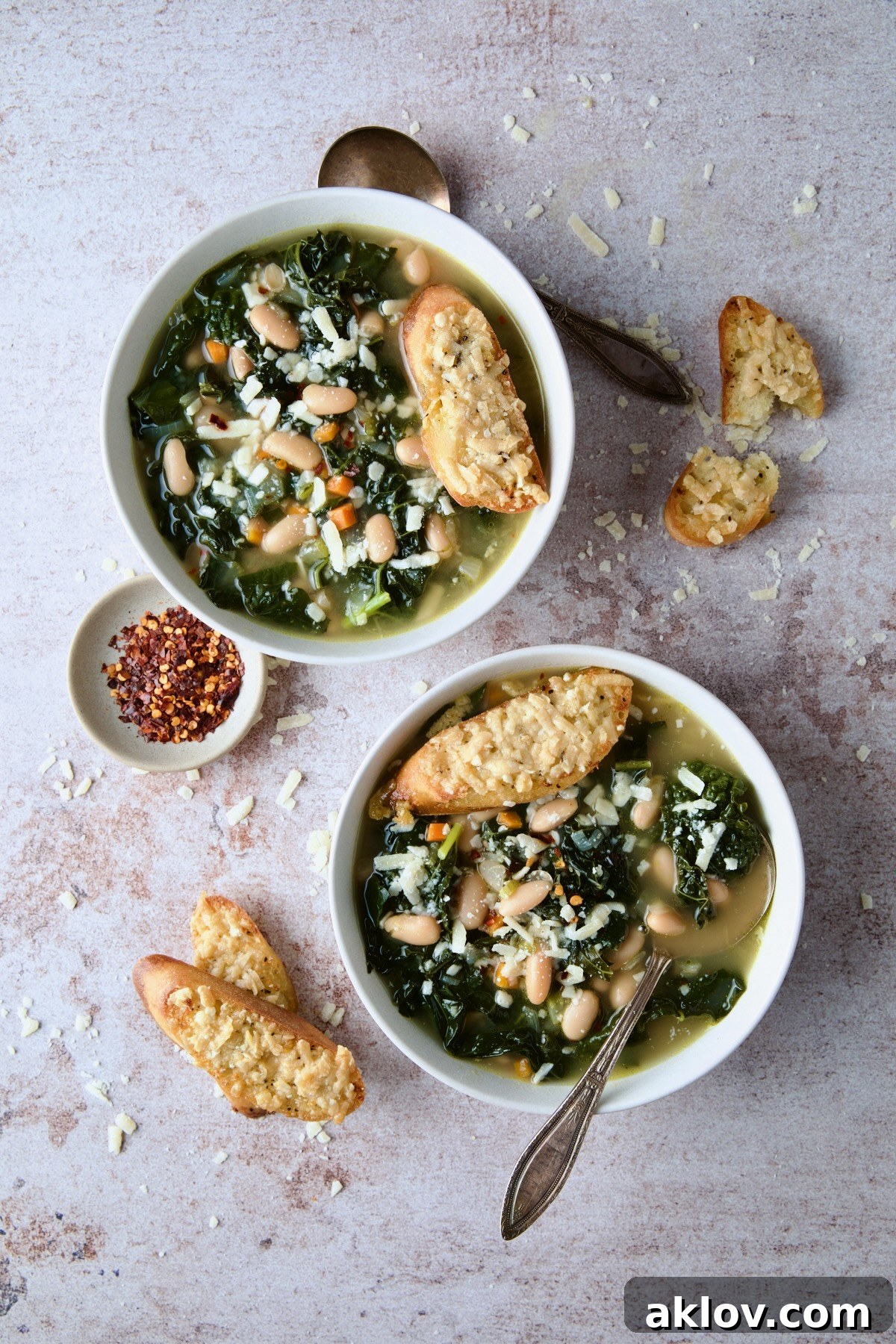 Two enticing bowls of White Bean Kale Soup, beautifully garnished with Parmesan Toast, sit alongside a small bowl of crushed red pepper flakes, inviting a customizable spice kick.