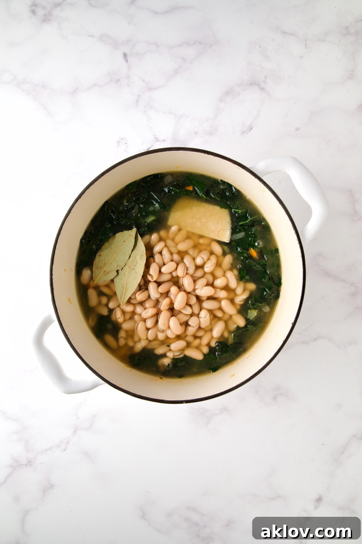 A view inside the Dutch oven shows the soup simmering gently with white beans, bay leaves, and a large piece of Parmigiano-Reggiano rind infusing flavor.