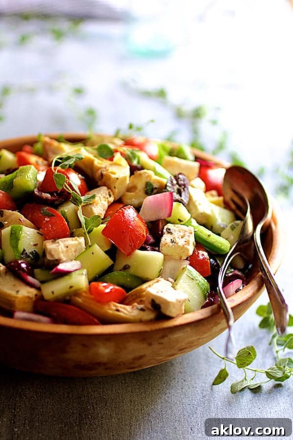 Photo of Greek Vegetable Salad with Marinated Feta Cheese in wood bowl with serving utensils garnished with fresh oregano.